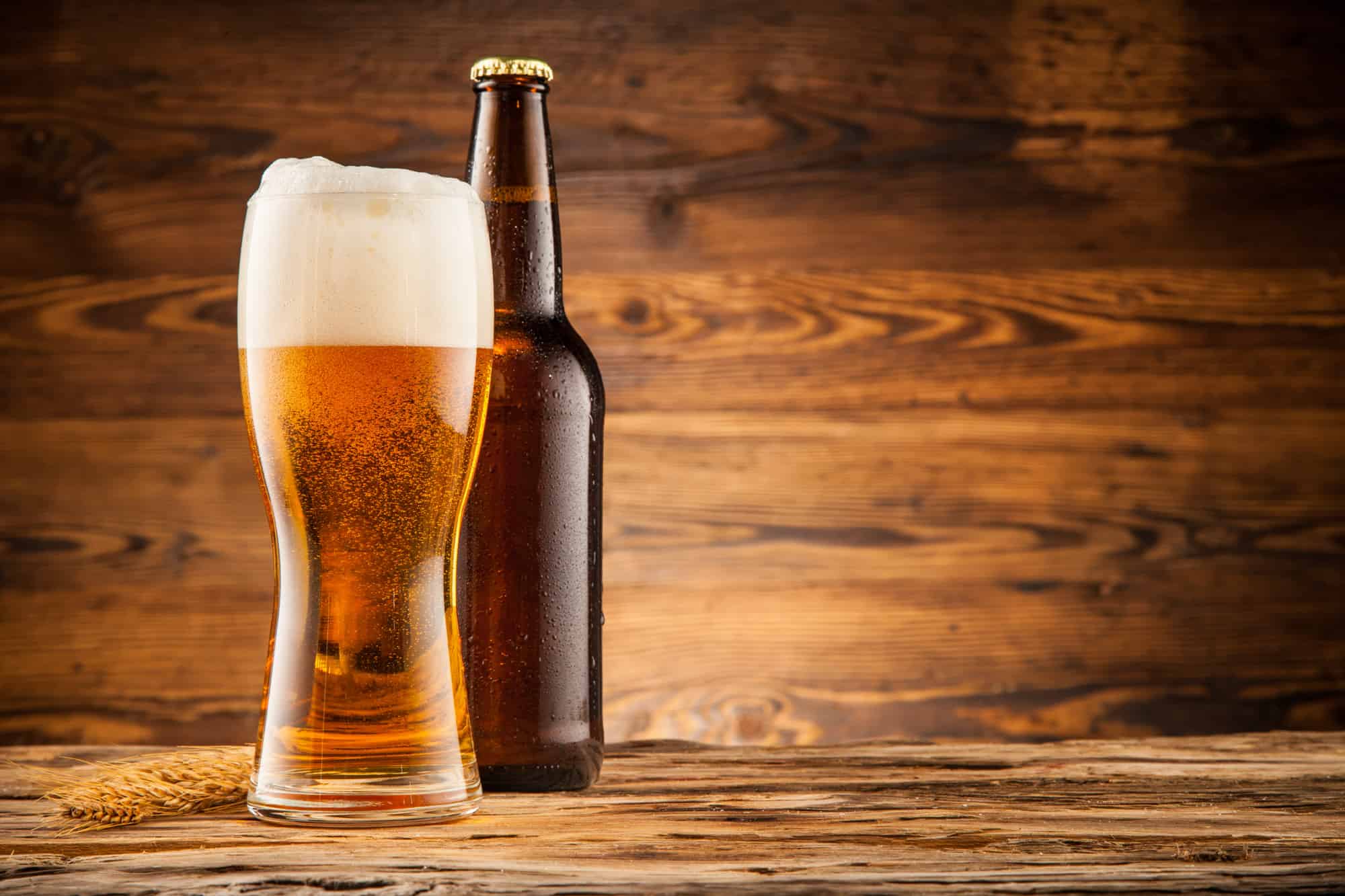 a tulip pint glass of beer with foam stands next to a bottle of beer in front of a wood background, is beer an acquired taste, yes and it takes some time to get used to beers like this one