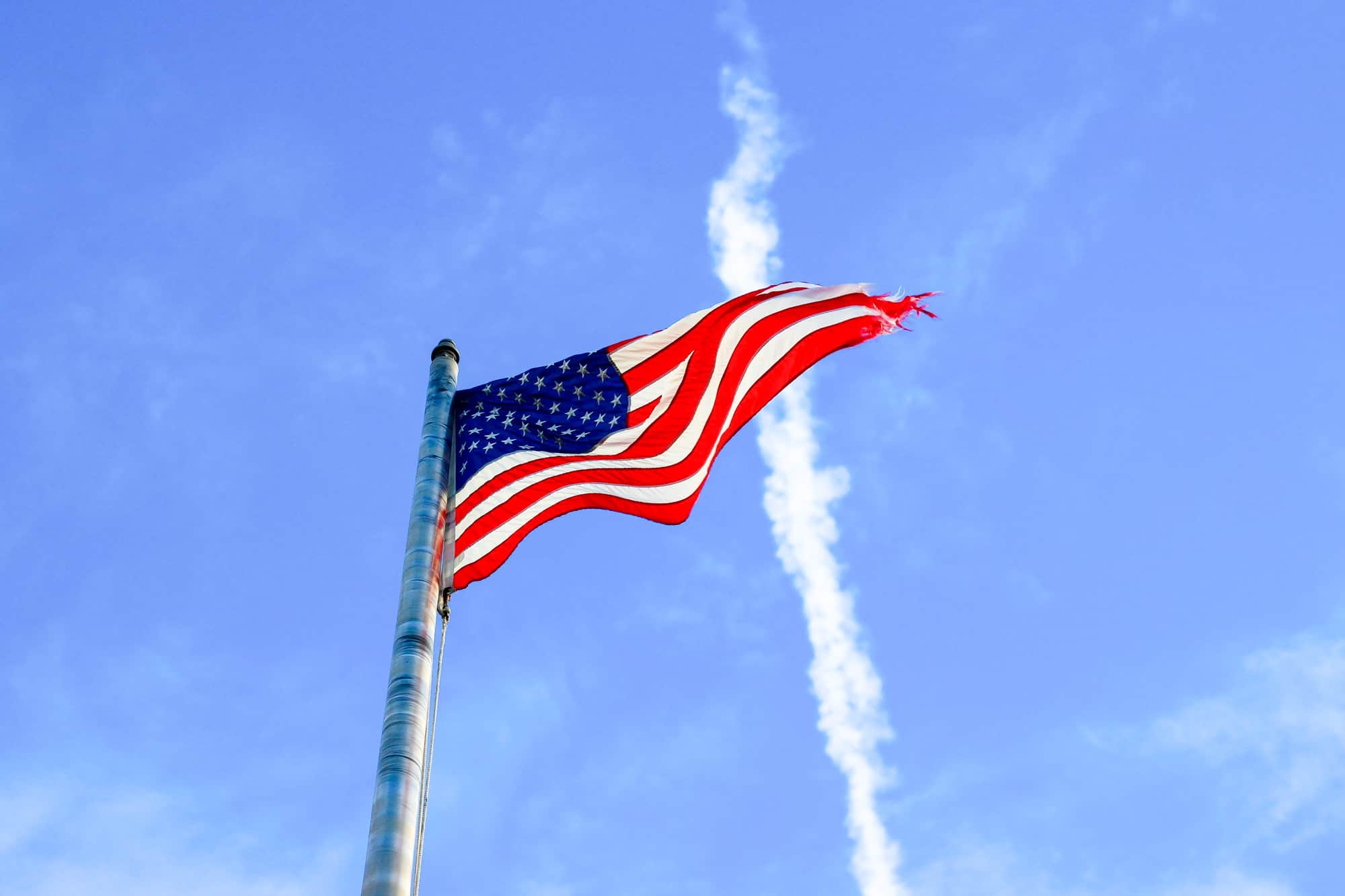an american flag with tattered ends flies at the top of a pole in front of a blue sky 