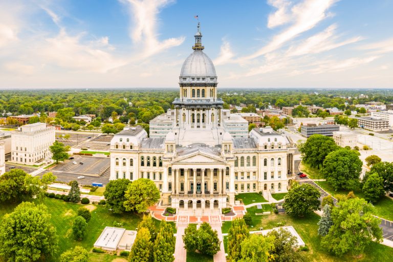a drone shot of the capitol building creating a birds eye view, the entire capitol can be seen with plenty of green trees on the grounds, under a blue sky