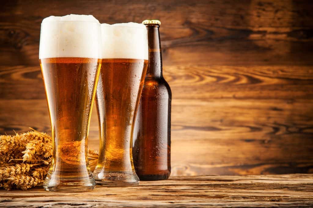 2 tall glasses of beer sit next to a dark beer bottle slighly off-center on the left. Lying next to the beer is some wheat stalks.